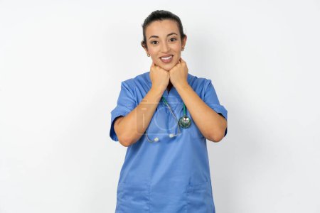 Foto de Satisfecho árabe médico mujer vistiendo azul uniforme toca la barbilla con ambas manos, sonríe agradablemente, se regocija buen día con el amante - Imagen libre de derechos