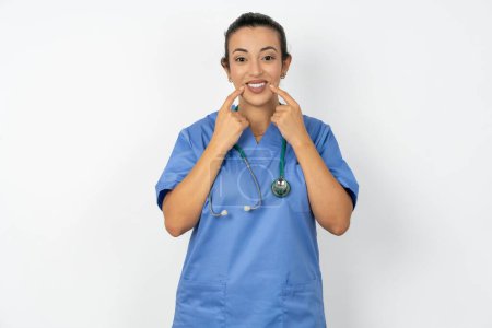 Téléchargez les photos : Médecin arabe femme portant un uniforme bleu avec sourire aux dents, garde les index près de la bouche, les doigts pointant et forçant sourire joyeux - en image libre de droit