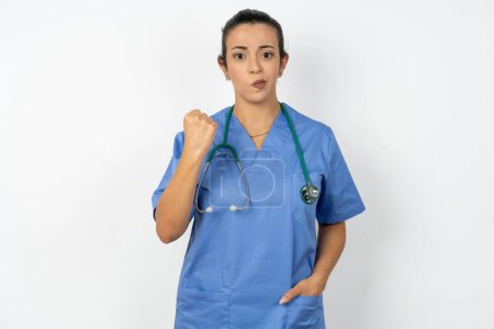 Photo for Arab doctor woman wearing blue uniform shows fist has annoyed face expression going to revenge or threaten someone makes serious look. I will show you who is boss - Royalty Free Image
