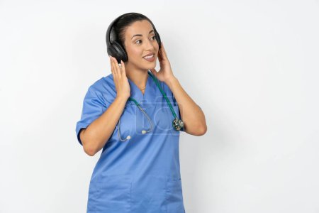 Photo for Arab doctor woman wearing blue uniform wears stereo headphones listens music concentrated aside. People hobby lifestyle concept - Royalty Free Image