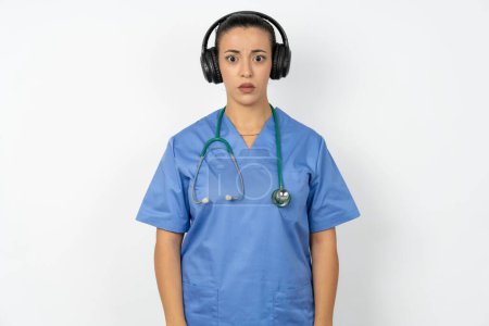 Téléchargez les photos : Grave mécontent médecin arabe femme portant uniforme bleu regarde perplexe à la caméra étant en colère porte casque stéréo écoute de la musique - en image libre de droit