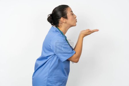 Téléchargez les photos : Vue de profil latérale, portrait d'une jolie médecin arabe femme portant un uniforme bleu envoyant un baiser d'air - en image libre de droit