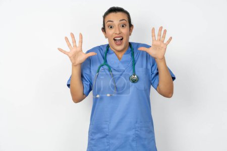 Téléchargez les photos : Délicieuse médecin arabe positive femme portant un uniforme bleu ouvre la bouche et les bras paumes vers le haut après avoir obtenu un excellent résultat - en image libre de droit