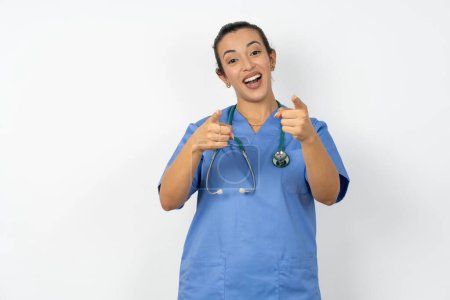 Foto de Árabe médico mujer vistiendo azul uniforme alegre y sonriente poiting a cámara - Imagen libre de derechos