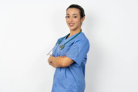 Foto de Retrato de mujer médico árabe vistiendo uniforme azul de pie con los brazos cruzados y sonriendo - Imagen libre de derechos