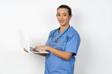 Téléchargez les photos : Adorable médecin arabe femme portant uniforme bleu tenant ordinateur portable - en image libre de droit