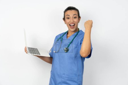 Téléchargez les photos : Arabe médecin femme portant uniforme bleu tenir ordinateur bouche ouverte lever poing - en image libre de droit