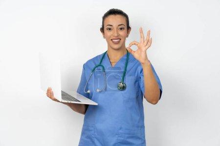 Foto de Positivo árabe doctor mujer usando azul uniforme hold inalámbrico netbook mano dedos mostrar okey símbolo - Imagen libre de derechos