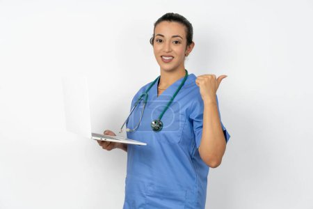 Photo for Arab doctor woman wearing blue uniform with laptop pointing empty space modern device - Royalty Free Image