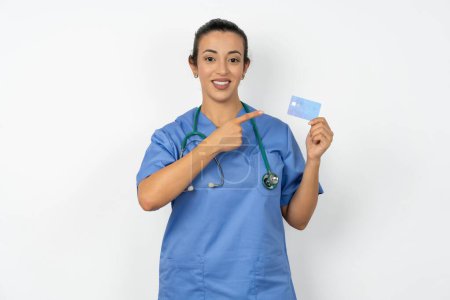 Téléchargez les photos : Portrait photo d'une femme médecin arabe portant un uniforme bleu faisant l'achat avec une carte bancaire pointant du doigt - en image libre de droit