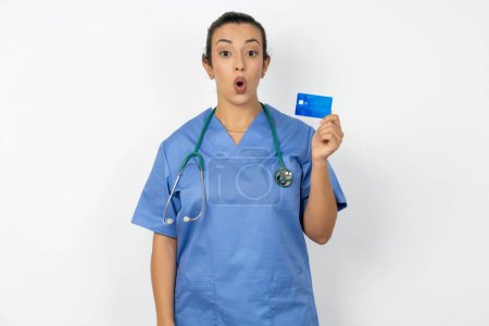 Foto de Foto de la mujer médico árabe vistiendo uniforme azul sorprendido celebrar pago con tarjeta de crédito - Imagen libre de derechos