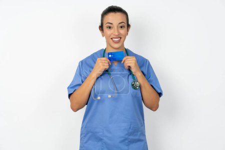 Foto de Foto de árabe médico mujer vistiendo azul uniforme sonrisa positiva mantener sueldo de la tarjeta de crédito - Imagen libre de derechos