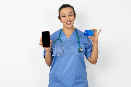 Téléchargez les photos : Médecin arabe femme portant uniforme bleu tenant smartphone et carte de crédit, souriant, recommander l'utilisation de l'application de magasinage en ligne - en image libre de droit