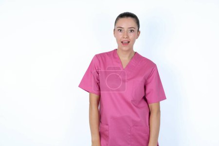 Téléchargez les photos : Femme caucasienne médecin en uniforme rose ayant regard stupéfait et choqué, avec la bouche ouverte et la mâchoire tombée en s'écriant : Wow, je ne peux pas croire cela. Surprise et choc - en image libre de droit