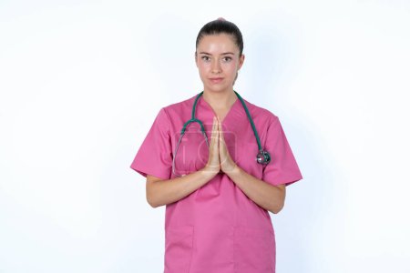 Photo for Caucasian woman doctor in pink uniform with stethoscope keeps palms pressed together in front of her having regretful look, asking for forgiveness. Forgive me please. - Royalty Free Image