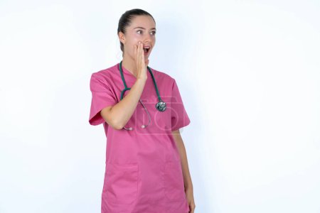 Foto de Médico mujer caucásica en uniforme rosa con vista de perfil estetoscopio, mirando feliz y emocionado, gritando y llamando para copiar espacio. - Imagen libre de derechos
