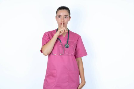 Téléchargez les photos : Femme caucasienne médecin en uniforme rose avec stéthoscope fait geste de silence, demande être calme. Ne dites pas mon secret ou ne parlez pas trop fort, s'il vous plaît.! - en image libre de droit