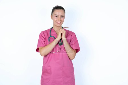 Photo for Dreamy caucasian woman doctor in pink uniform with stethoscope with pleasant expression, keeps hands crossed near face, excited about something pleasant. - Royalty Free Image
