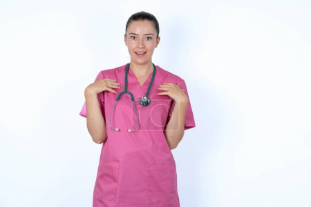 Photo for Caucasian woman doctor in pink uniform with stethoscope keeps hands on chest feeling shocked and scared, mouth widely opened, stares at camera saying: Who, me? - Royalty Free Image