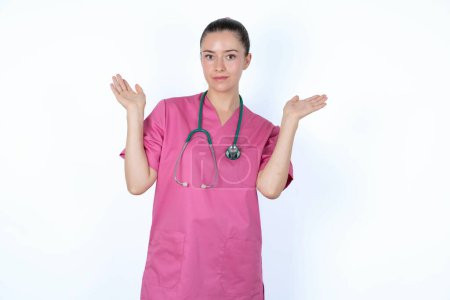 Téléchargez les photos : Femme caucasienne perplexe et désemparée médecin en uniforme rose avec stéthoscope les bras ouverts, haussant les épaules, disant : qui s'en soucie, alors quoi, je ne sais pas. - en image libre de droit