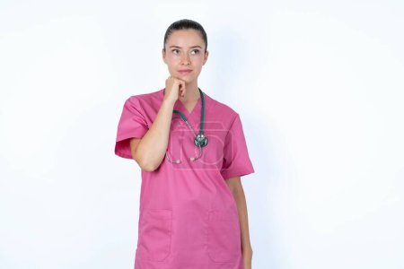 Photo for Portrait of thoughtful  caucasian woman doctor in pink uniform with stethoscope  keeps hand under chin, looks away trying to remember something or listens something with interest. Youth concept. - Royalty Free Image