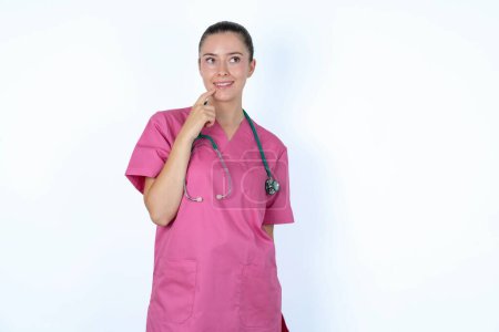 Photo for Caucasian woman doctor in pink uniform with stethoscope with thoughtful expression, looks to the camera, keeps hand near face, bitting a finger thinks about something pleasant. - Royalty Free Image