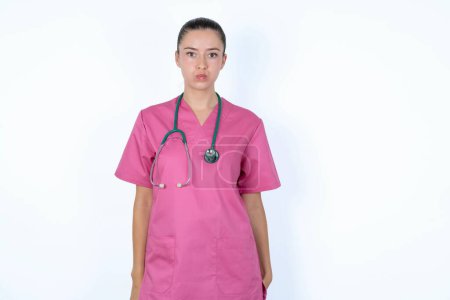 Téléchargez les photos : Femme caucasienne médecin en uniforme rose avec stéthoscope joues gonflées avec visage drôle. Bouche gonflée avec de l'air, expression folle. - en image libre de droit