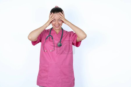 Foto de Doctora caucásica conmocionada en uniforme rosa con estetoscopio agarrado de la cabeza y gritando en desesperación y frustración. - Imagen libre de derechos