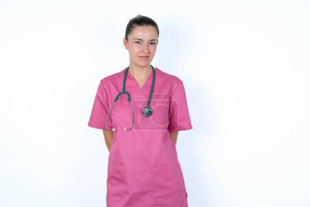 Photo for Caucasian woman doctor in pink uniform with stethoscope clenches teeth angrily, being annoyed with coming noise. Negative feeling concept. - Royalty Free Image
