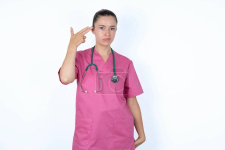 Foto de Mujer caucásica infeliz médico en uniforme rosa con estetoscopio imita disparo de arma hace gesto suicida mantiene dos dedos en las sienes. - Imagen libre de derechos
