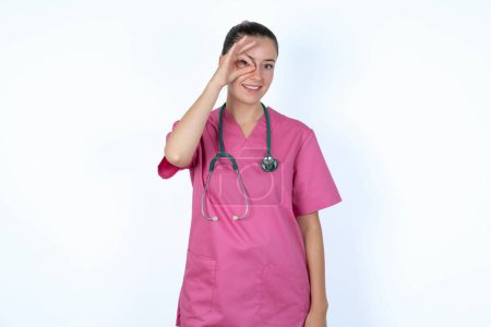 Téléchargez les photos : Femme caucasienne médecin en uniforme rose avec stéthoscope avec le visage heureux souriant faire ok signe avec la main sur l'oeil regardant à travers le doigt. - en image libre de droit