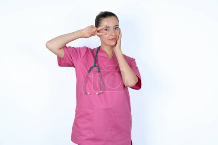Téléchargez les photos : Femme médecin caucasienne en uniforme rose avec stéthoscope faisant signe en V près des yeux. Loisirs style de vie personne célébrer flirt concept coquette. - en image libre de droit