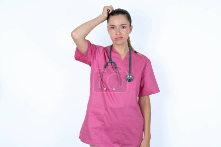 Téléchargez les photos : Femme médecin caucasienne en uniforme rose avec stéthoscope disant : Oups, qu'est-ce que j'ai fait ? Tenant la main sur la tête avec une expression effrayée et de regret. - en image libre de droit