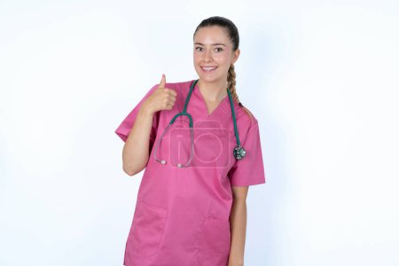 Foto de Médico mujer caucásica en uniforme rosa con estetoscopio dando el pulgar hacia arriba gesto, buen trabajo! Emoción humana positiva expresión facial lenguaje corporal. - Imagen libre de derechos