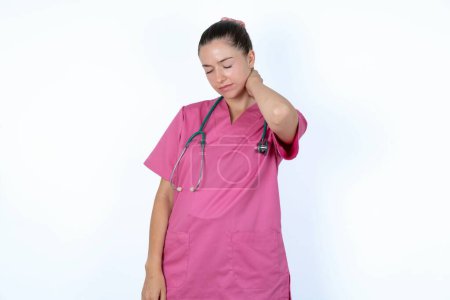 Photo for Caucasian woman doctor in pink uniform with stethoscope suffering from back and neck ache injury, touching neck with hand, muscular pain. - Royalty Free Image