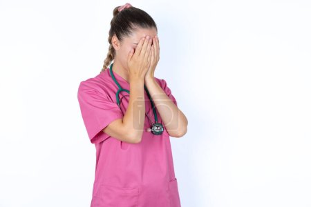 Téléchargez les photos : Triste médecin caucasienne en uniforme rose avec stéthoscope couvrant le visage avec les mains et pleurant. - en image libre de droit