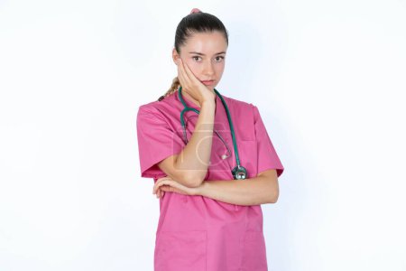 Photo for Very bored caucasian female doctor in pink uniform with stethoscope holding hand on cheek while support it with another crossed hand, looking tired and sick. - Royalty Free Image