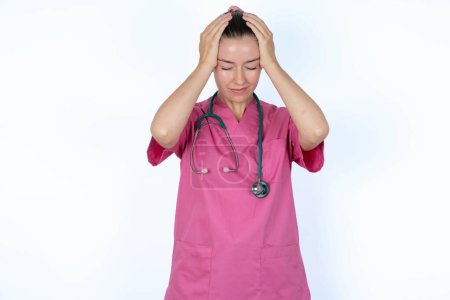 Foto de Médico femenino caucásico en uniforme rosa con estetoscopio sosteniendo la cabeza con las manos, sufriendo de dolor de cabeza severo, presionando los dedos a las sienes - Imagen libre de derechos