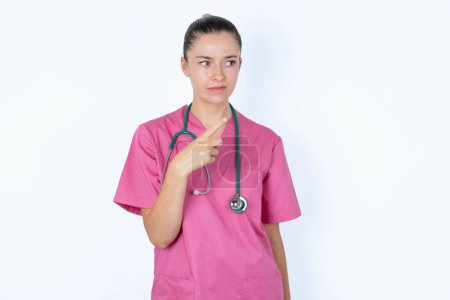 Téléchargez les photos : Heureuse femme caucasienne médecin regardant et pointant de côté avec la main. Espace de copie et concept publicitaire. - en image libre de droit