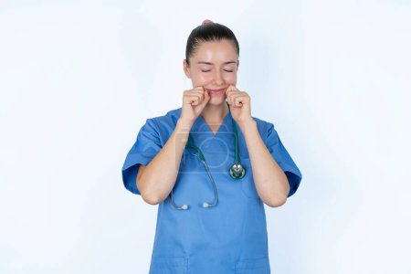 Photo for Pleased caucasian female doctor with closed eyes keeps hands near cheeks and smiles tenderly imagines something very pleasant - Royalty Free Image