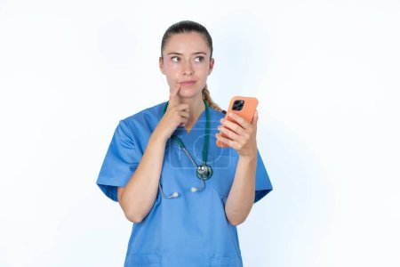 Photo for Caucasian female doctor thinks deeply about something, uses modern mobile phone, tries to made up good message, keeps index finger near lips. - Royalty Free Image