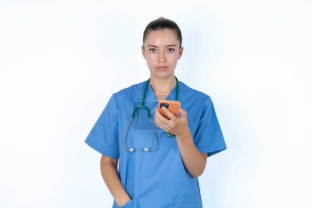 Foto de Molesto médico femenino insatisfecho utiliza aplicación de software móvil y navega información en Internet, sostiene la mano móvil moderna - Imagen libre de derechos