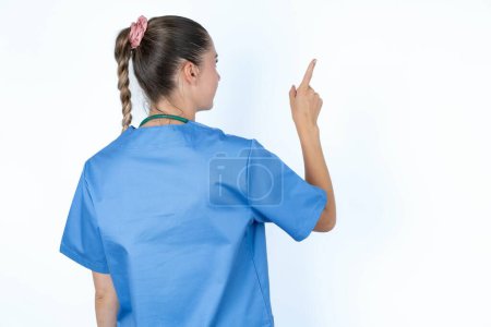 Téléchargez les photos : Femme médecin caucasienne en uniforme avec stéthoscope pointant vers l'objet sur l'espace de copie, vue arrière. Tourne le dos - en image libre de droit