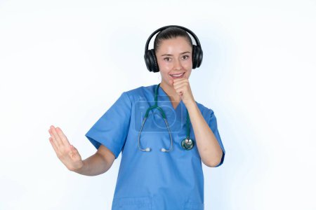 Foto de Mujer caucásica médico en uniforme con estetoscopio canta canción favorita mantiene la mano cerca de la boca como si el micrófono lleva auriculares inalámbricos, escucha música - Imagen libre de derechos