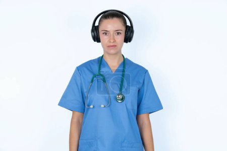 Téléchargez les photos : Portrait sérieux de femme caucasienne triste médecin en uniforme avec stéthoscope semble perplexe à la caméra étant en colère porte casque stéréo écoute de la musique - en image libre de droit
