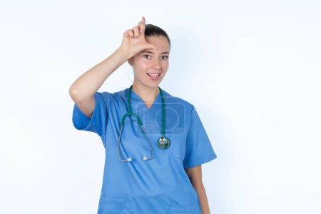Photo for Funny caucasian woman doctor in  uniform with stethoscope  makes loser gesture mocking at someone sticks out tongue making grimace face. - Royalty Free Image