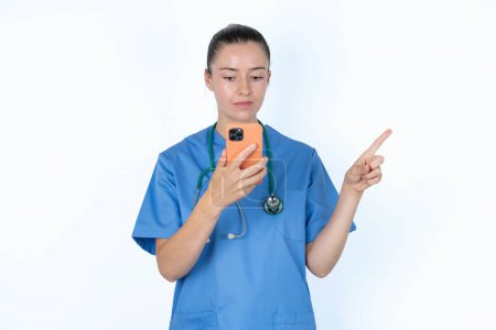Foto de Médico de mujer caucásica en uniforme con estetoscopio apuntando con el dedo al espacio en blanco sosteniendo el teléfono en una mano - Imagen libre de derechos