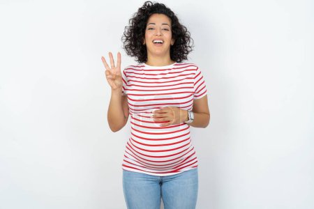 Foto de Mujer embarazada mostrando y apuntando hacia arriba con los dedos número tres mientras sonríe confiado y feliz. - Imagen libre de derechos