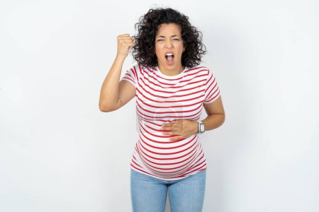 mujer embarazada enojada y loca levantando el puño frustrado y furioso mientras gritaba con ira. Rabia y concepto agresivo.