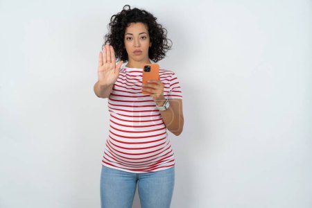 Foto de Mujer embarazada usando y mensajes de texto con teléfono inteligente con la mano abierta haciendo stop sign con expresión seria y segura, gesto de defensa - Imagen libre de derechos